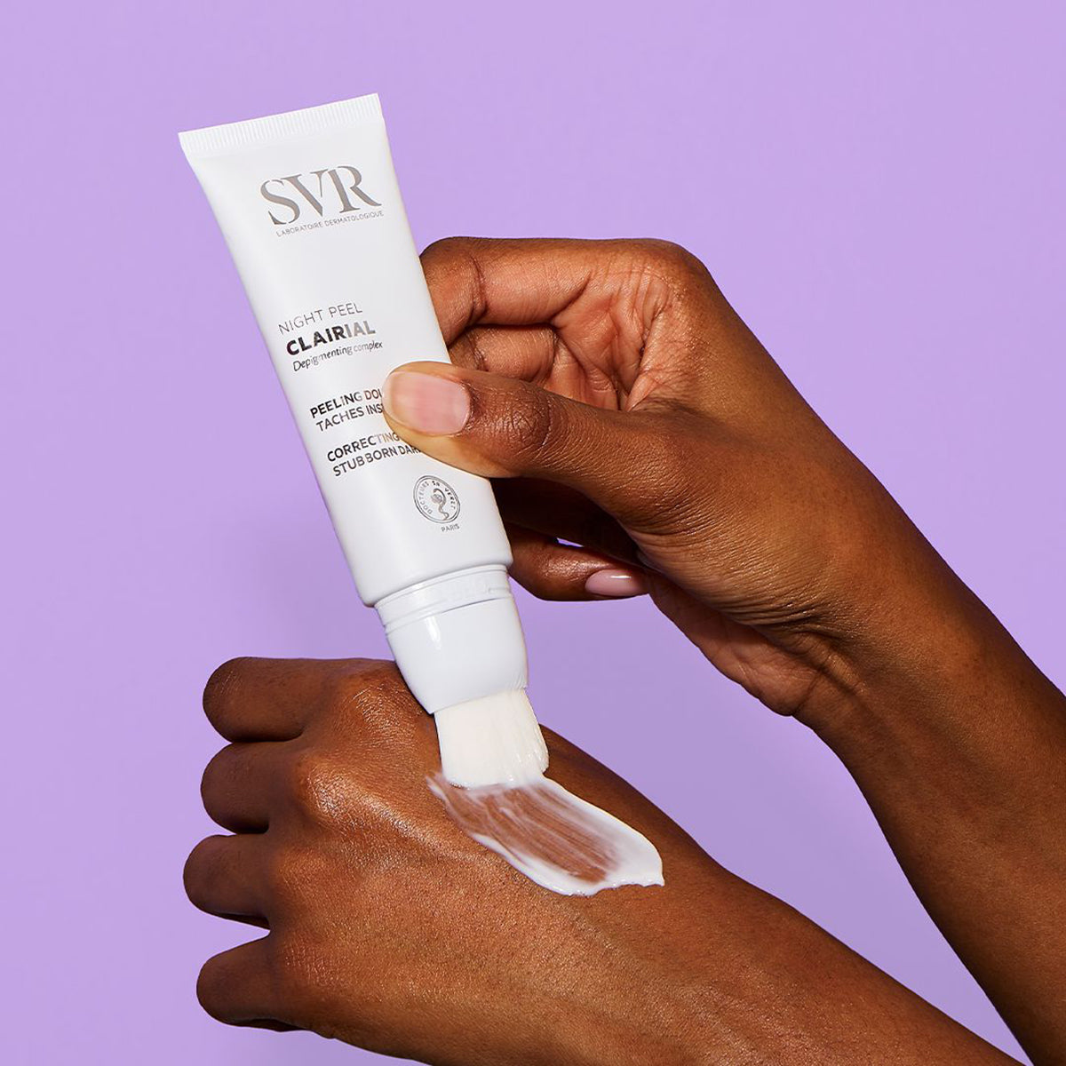 SVR Clairial Night Peel white tube with brush applicator applying cream to a hand against a purple background.