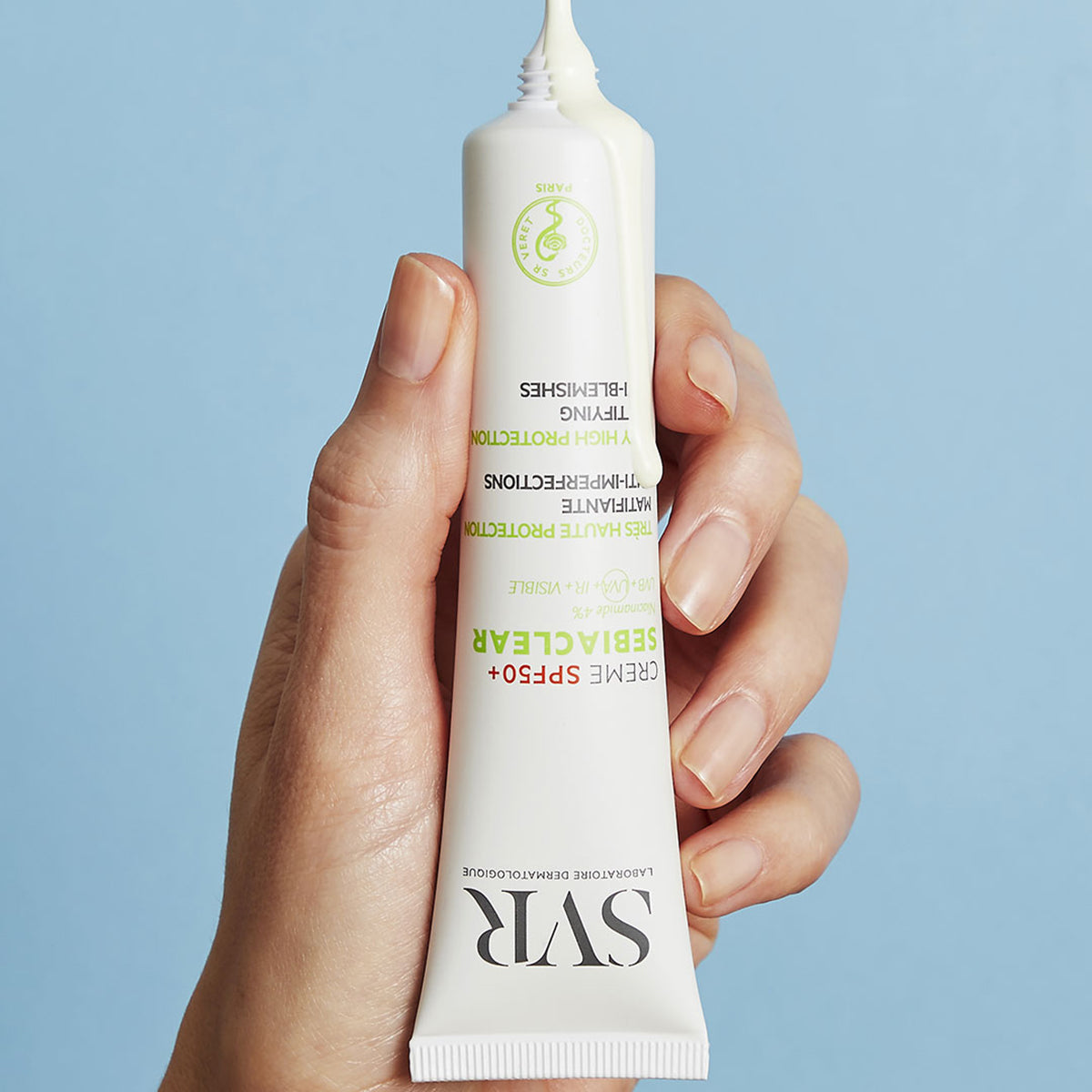 Hand holding a white SVR Sebiaclear SPF50+ cream tube with product dispensing from the top against a blue background.