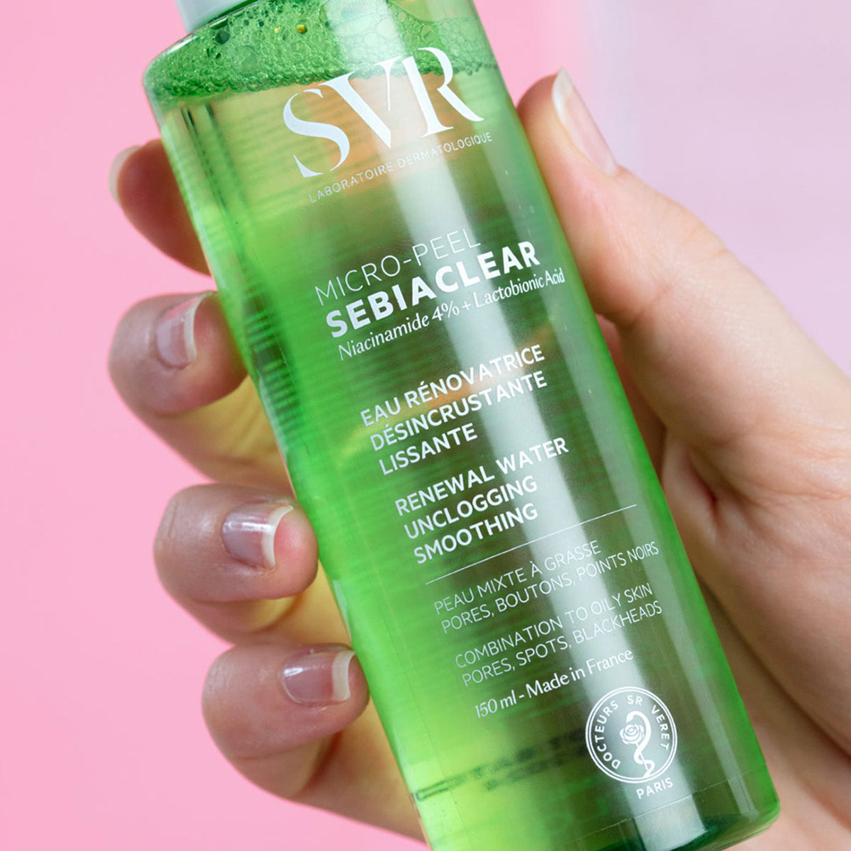 Hand holding a clear green 150ml bottle of SVR Sebiaclear Micro-Peel renewal water against a pink background.