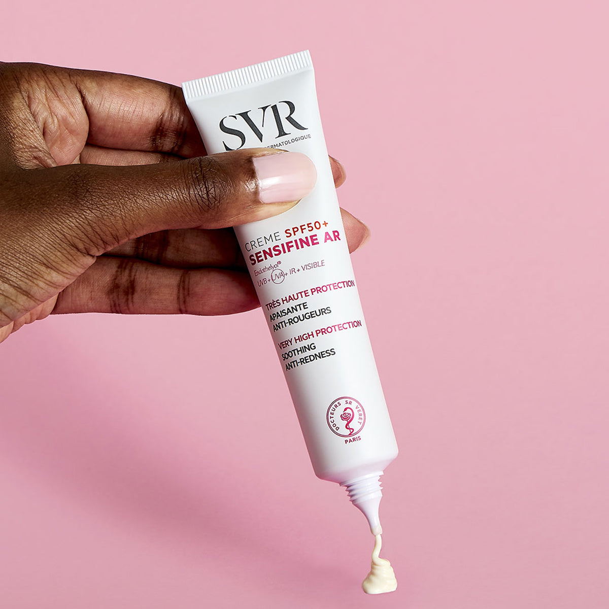 Hand holding a white SVR Sensifine AR SPF50+ cream tube with a drop of beige product against a pink background.