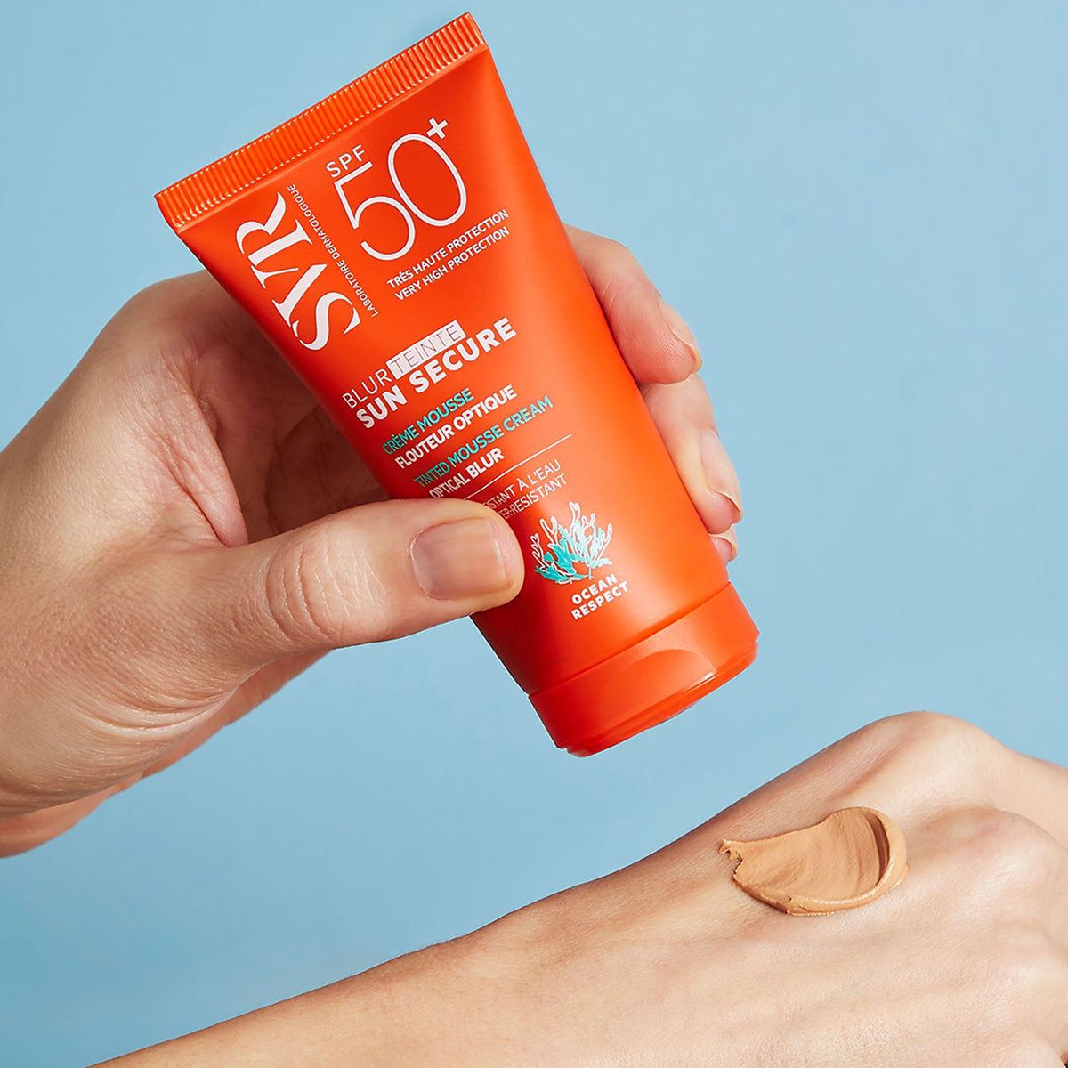 Hand holding an orange SVR Sun Secure SPF50 tube, applying a beige tinted mousse cream onto skin against a blue background.