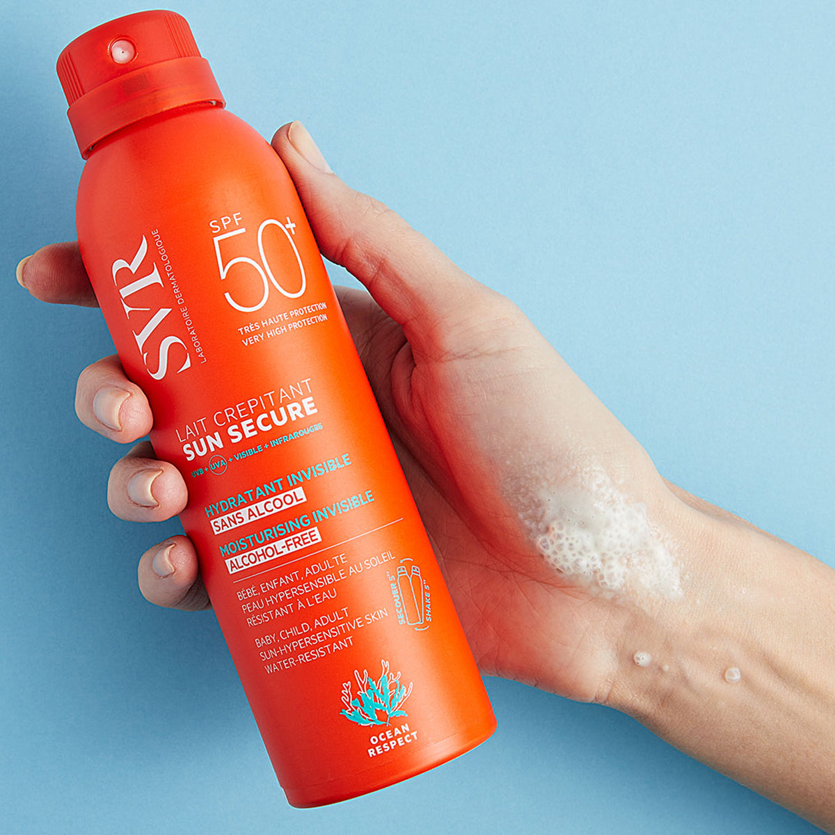 Hand holding an orange SVR SUN SECURE SPF50+ spray bottle with white foam texture on the skin against a blue background.
