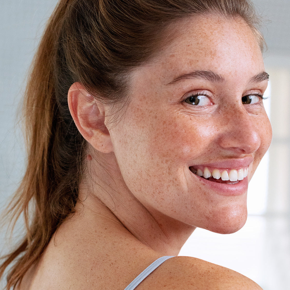 Smiling woman with freckles and natural skin texture looking over her shoulder, highlighting healthy, hydrated lips.