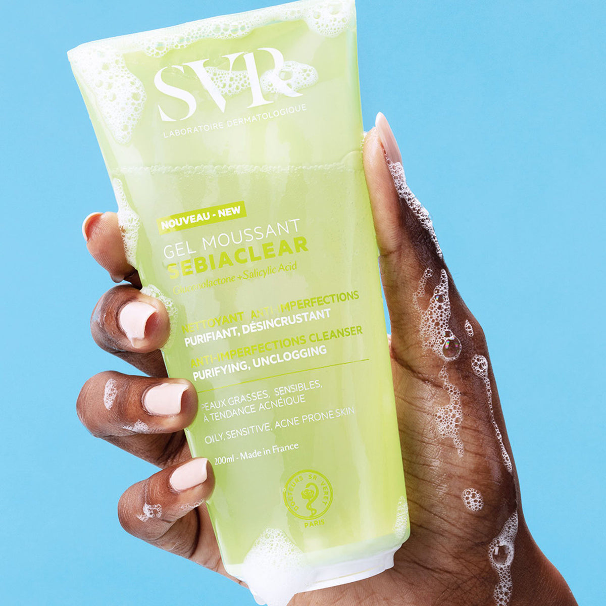 Hand holding a 200ml tube of SVR Sebiaclear Gel Moussant cleanser with soapy bubbles against a blue background.