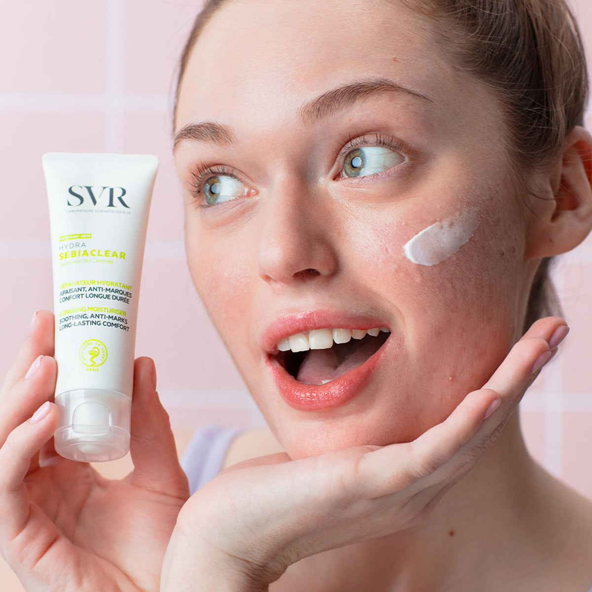 Woman holding a white SVR Sebiaclear Hydra moisturizer tube with a dab of cream on her cheek against a pink tiled background.