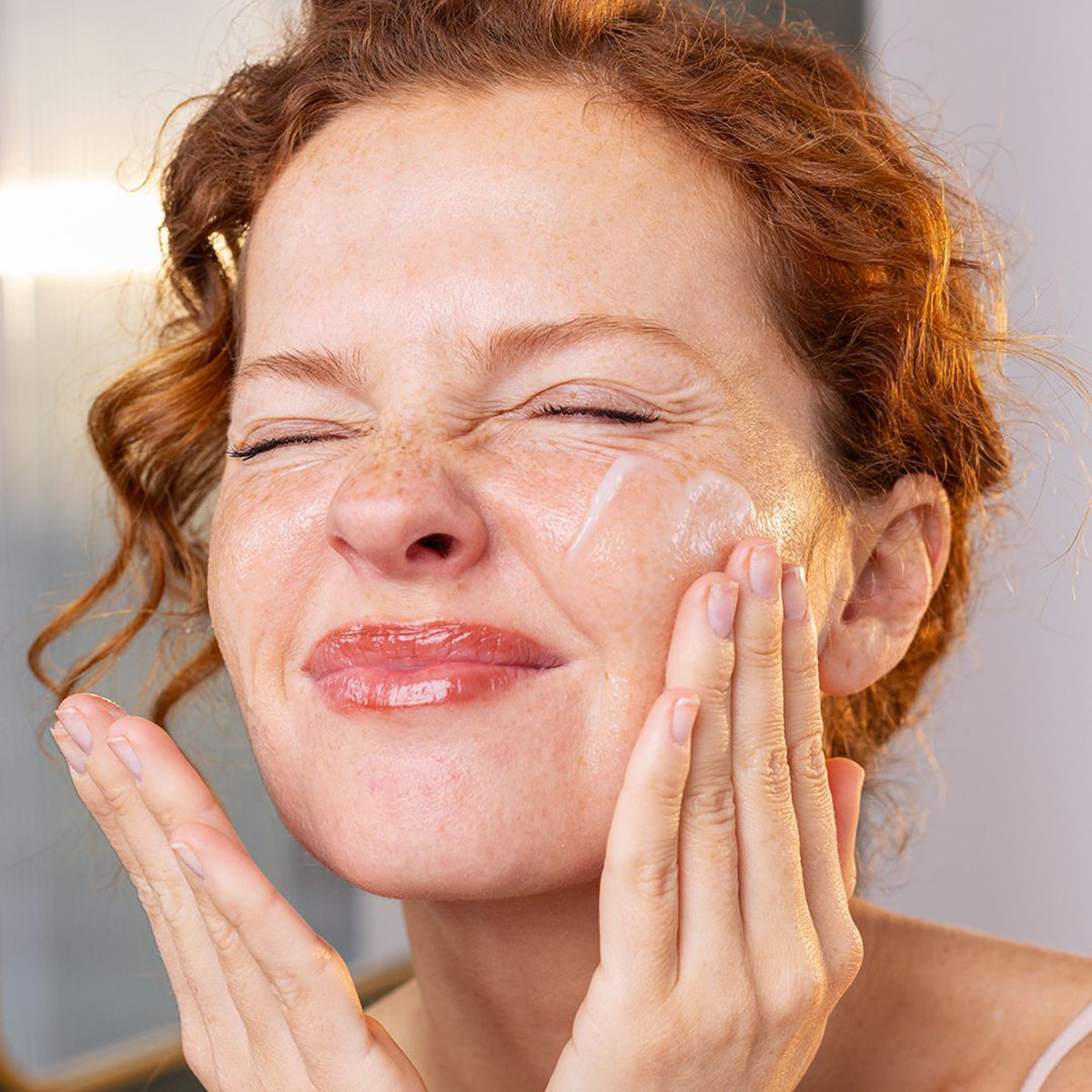 Woman with curly red hair applying white Sensifine Baume Demaquillant cleansing balm to her cheek.