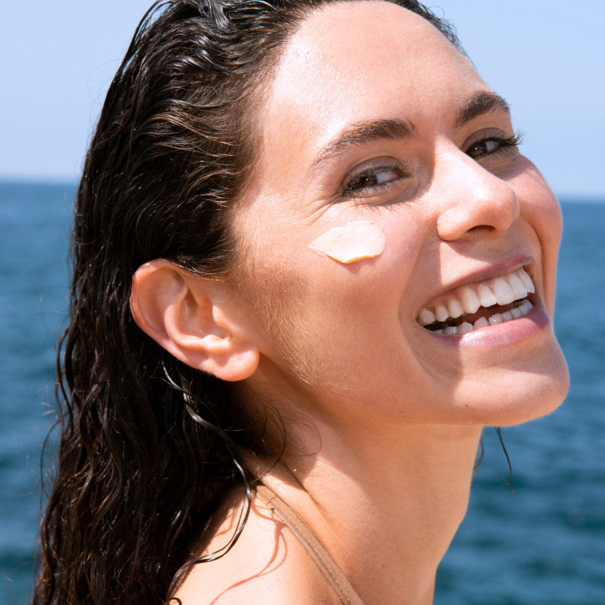 Smiling woman with wet hair applying beige SUN SECURE Blur SPF50+ cream on her cheek against a blue ocean background.