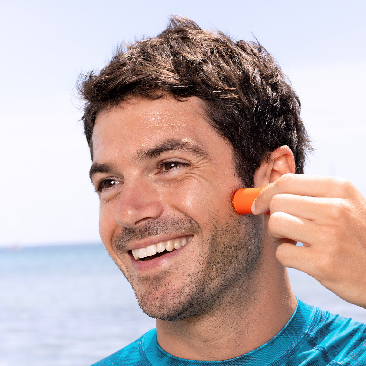 Smiling man applying orange SUN SECURE SPF50+ sunscreen stick to his cheek against a sunny beach background.