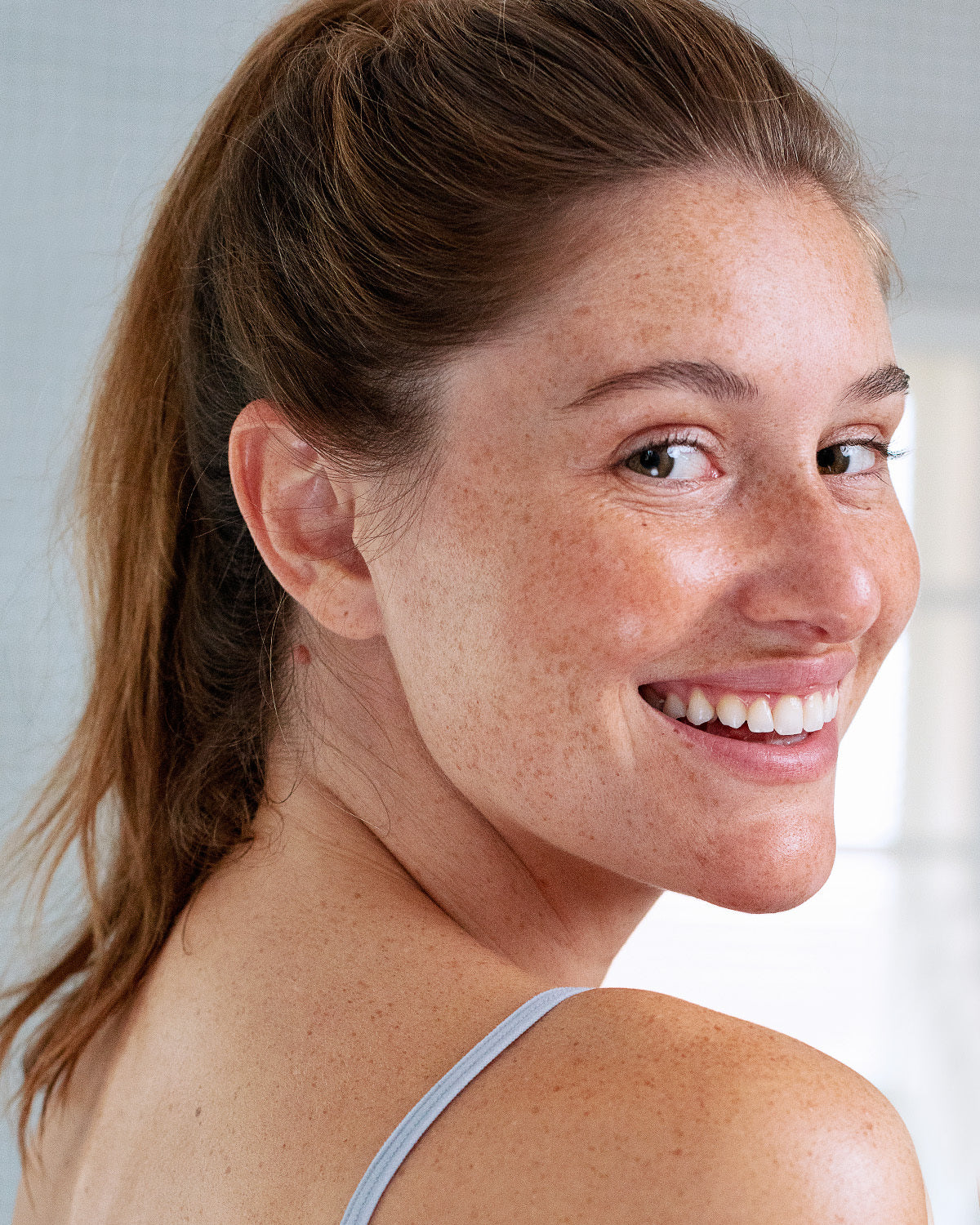 Smiling woman with freckles and brown hair in a ponytail, showing clear, healthy skin over her shoulder.