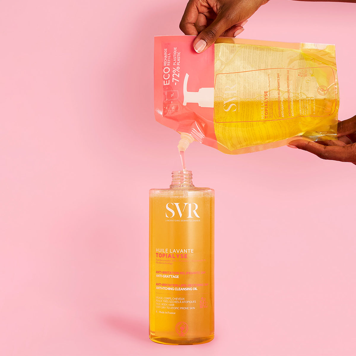 Hands pouring golden SVR TOPIALYSE Huile Lavante refill pouch into a clear plastic bottle against a pink background.