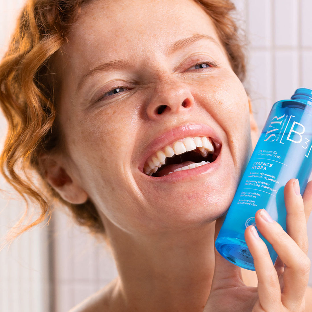 Smiling woman holding a blue SVR B3 Essence Hydra bottle for sensitive, dehydrated skin.