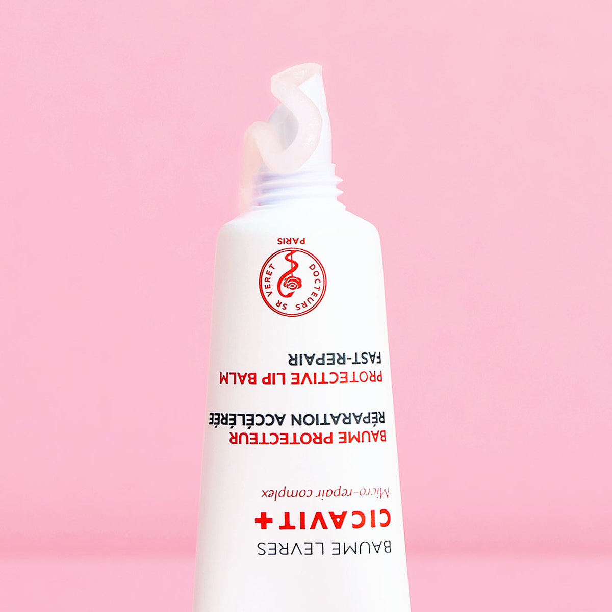 White squeeze tube of CICAVIT+ Lèvres protective lip balm with a dollop of clear gel on a pink background.