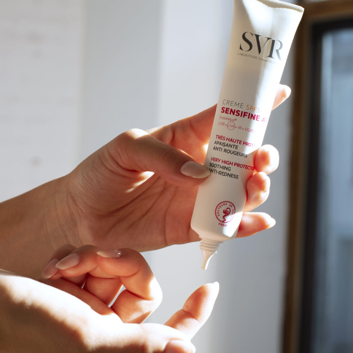 Hand holding a white tube of SVR Sensifine AR Creme SPF50+ with red text, dispensing a drop of cream onto a fingertip.