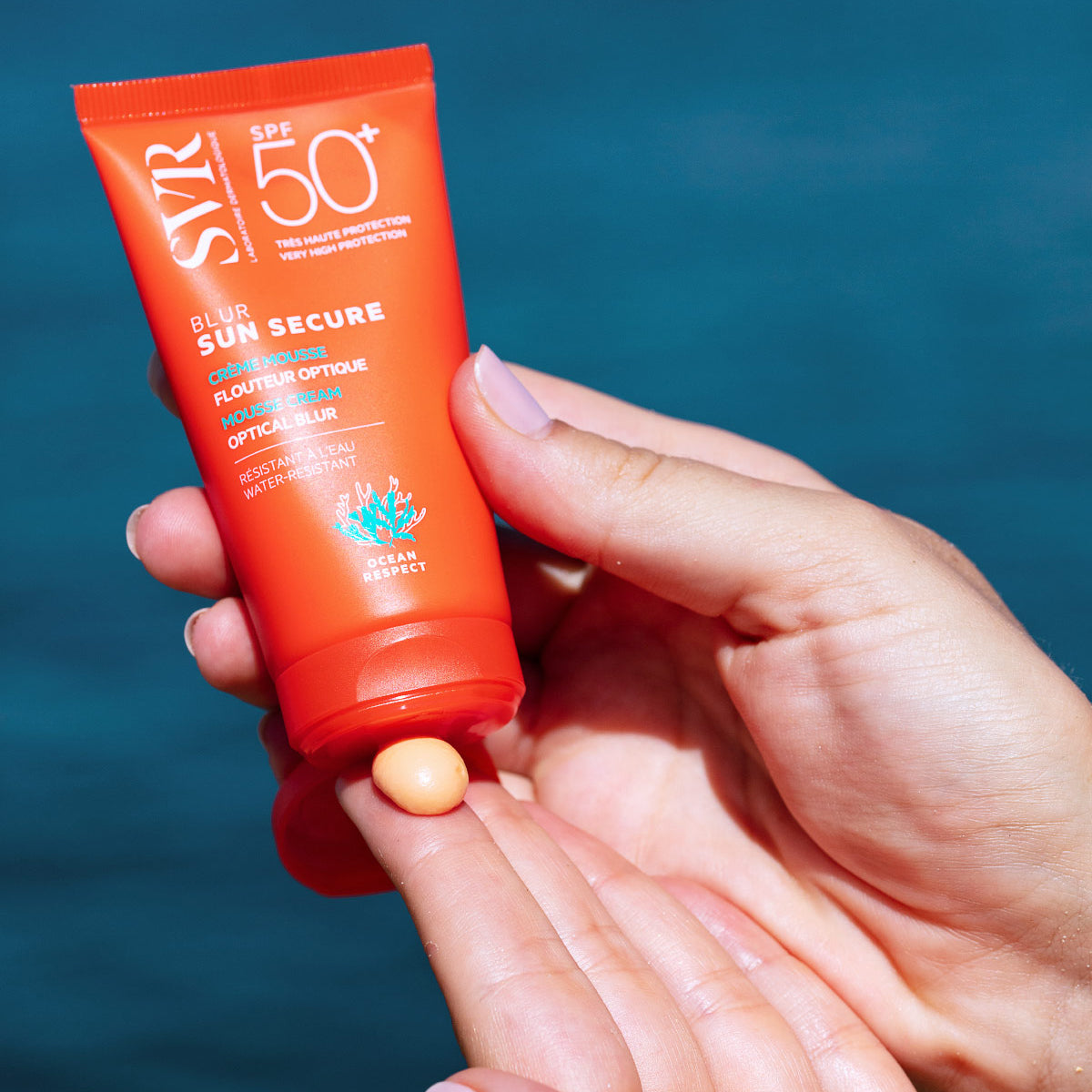 Orange SVR SUN SECURE Blur SPF50+ mousse cream tube being squeezed onto a finger against a blue water background.