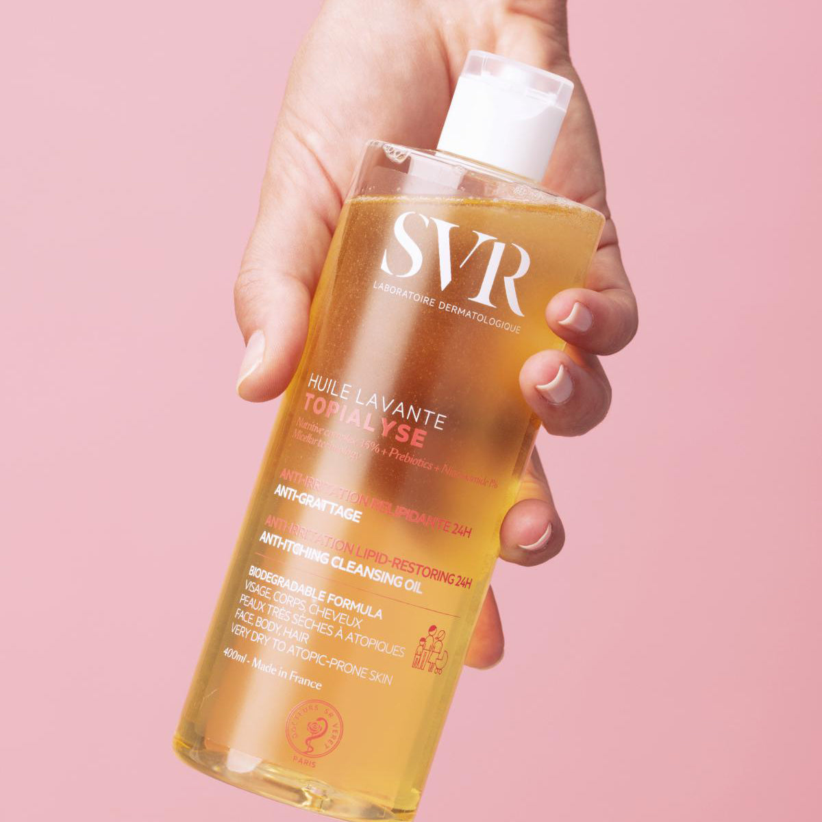 Hand holding a 400ml clear bottle of SVR Topialyse Huile Lavante cleansing oil against a soft pink background.