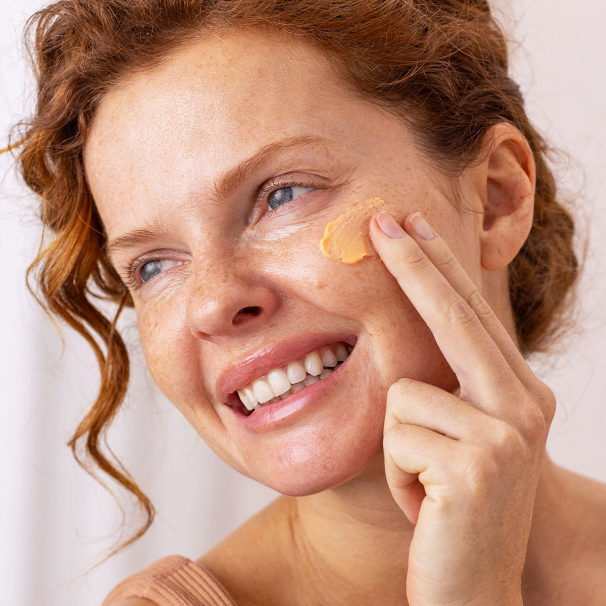 Smiling woman with red hair applying a peach-toned SUN SECURE Blur SPF50+ cream to her cheek.