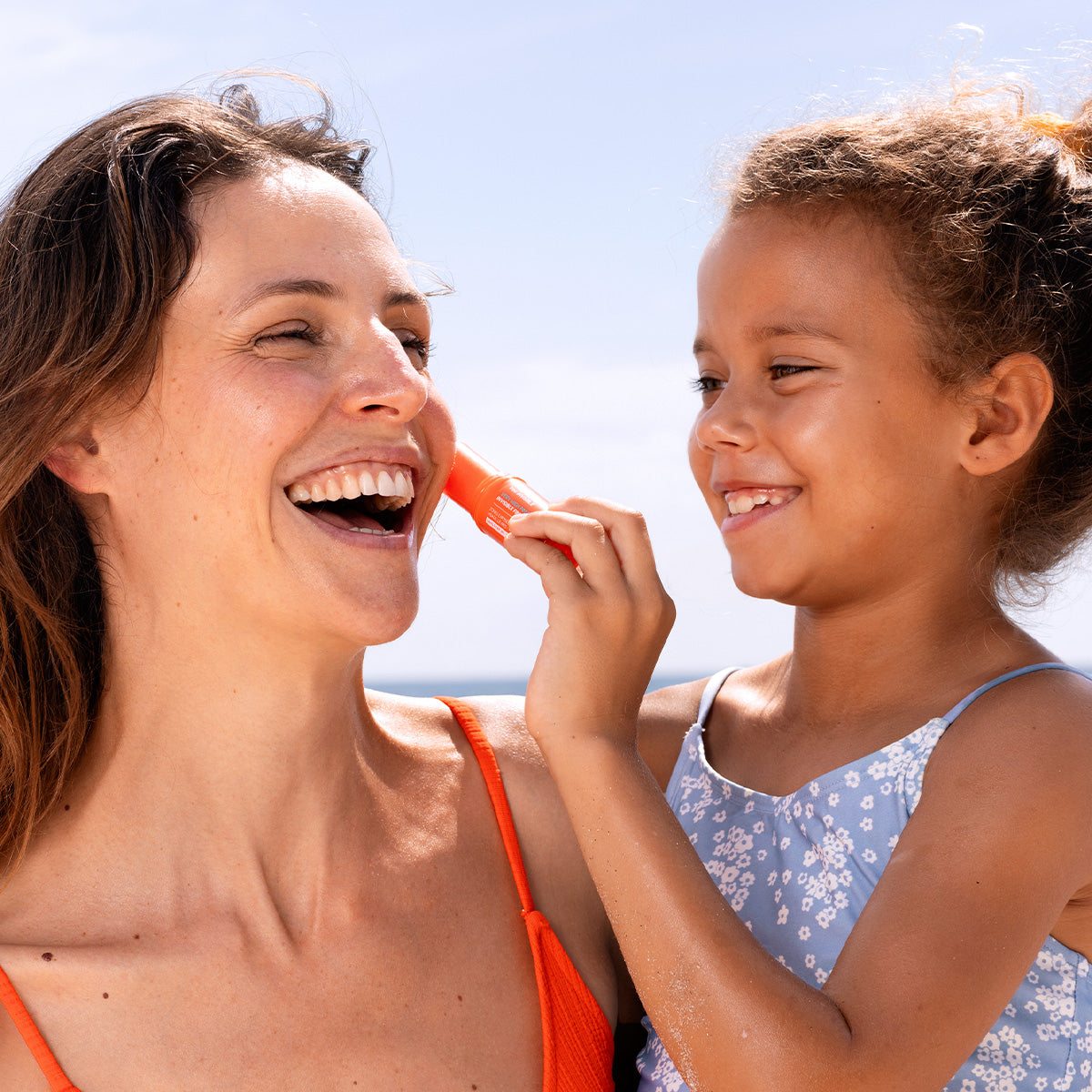 Young girl applying orange SUN SECURE Easy Stick SPF50+ sunscreen to her mother's cheek on a sunny beach day.