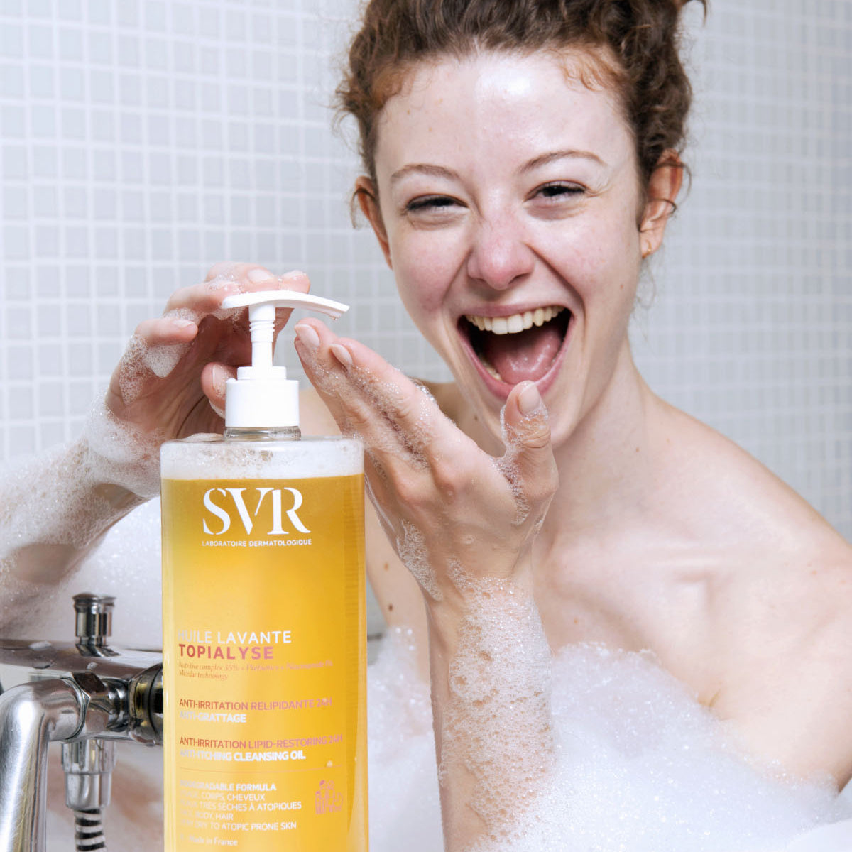 Smiling woman in a bubble bath holding a large, clear pump bottle of SVR TOPIALYSE Huile Lavante cleansing oil.