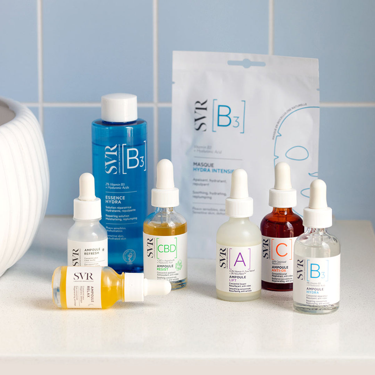SVR B3 Essence Hydra and various skincare ampoules with droppers arranged on a white surface against a blue tiled wall.