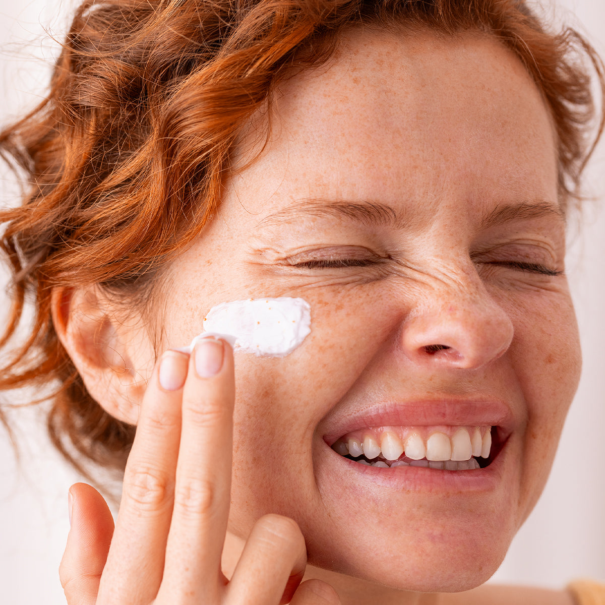 Smiling woman with red hair applying white C20 Biotic face cream to her cheek.