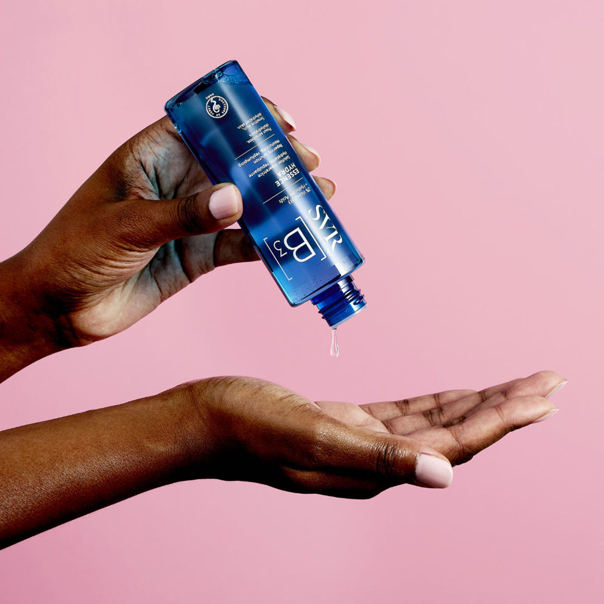 SVR B3 Essence Hydra blue bottle pouring a clear hydrating facial serum into a hand against a pink background.