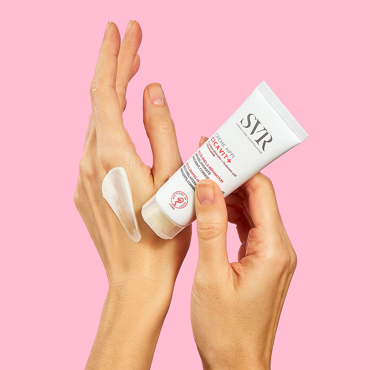 White SVR Cicavit+ Crème HPPI tube being applied to a hand, showing a smooth white cream texture against a pink background.