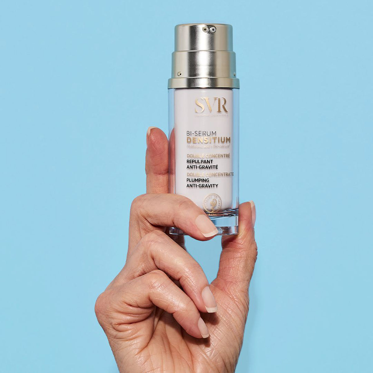 Hand holding a SVR Densitium Bi-Serum bottle with a gold pump cap against a light blue background.