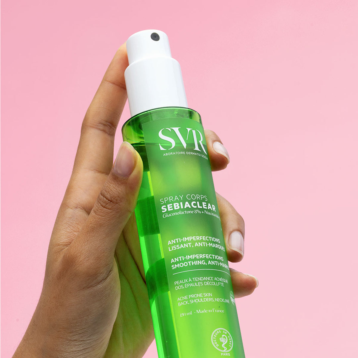 Hand holding a 150ml green transparent SVR Sebiaclear body spray bottle against a pink background.
