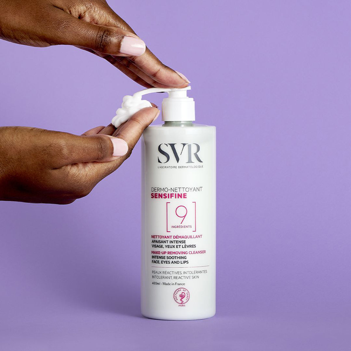 Hands dispensing white foam from a 400ml SVR Sensifine Dermo-Nettoyant cleanser bottle against a purple background.