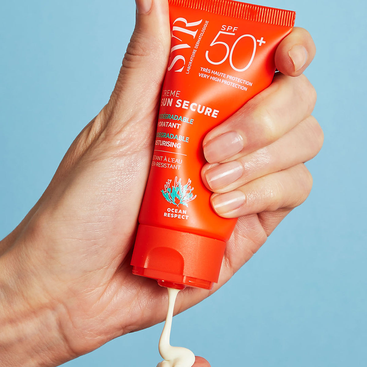 Hand squeezing an orange SVR SUN SECURE SPF50+ sunscreen tube, dispensing white cream against a blue background.
