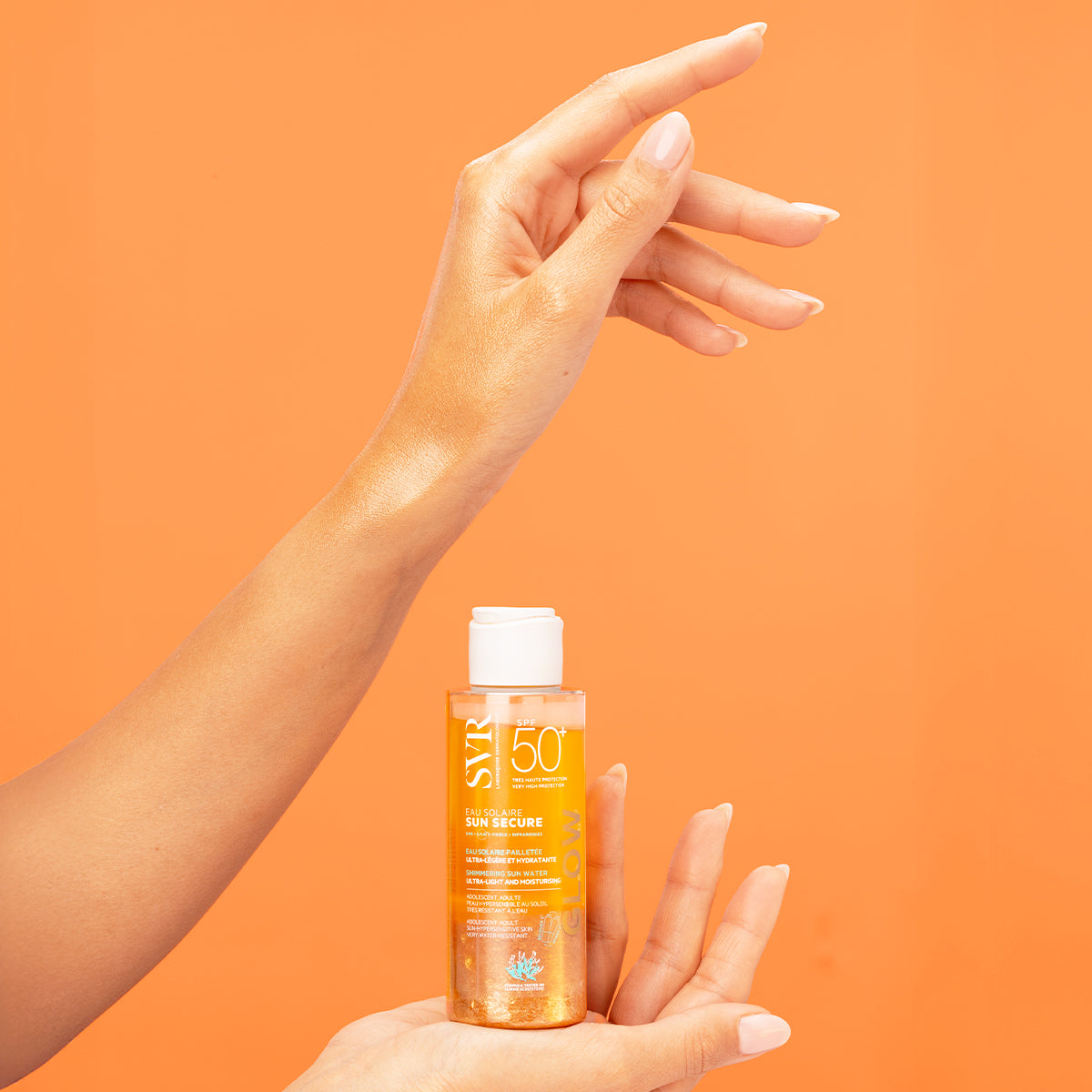 Hand holding a bottle of SVR SUN SECURE SPF50+ shimmering sun water against a bright orange background.