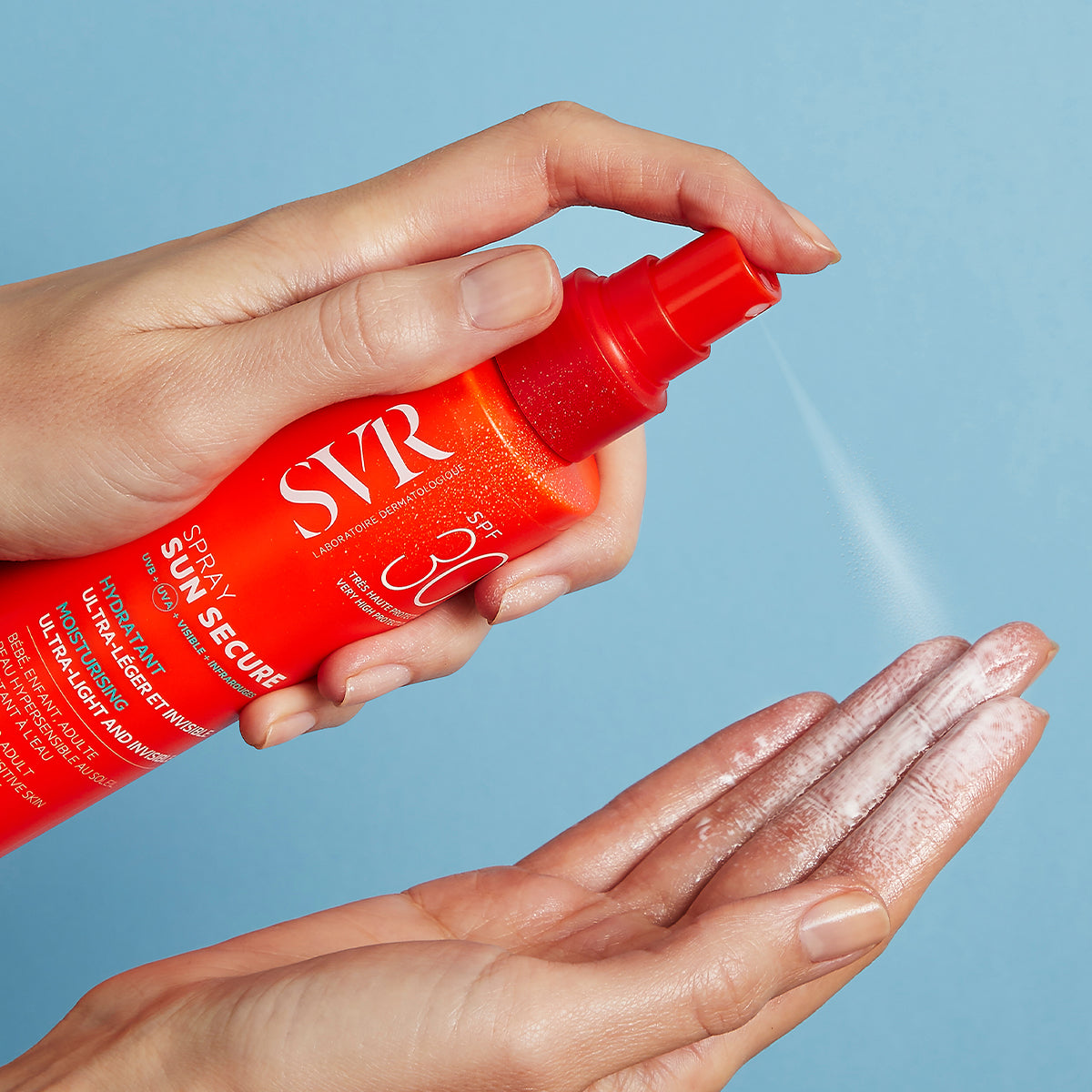 Hands spraying SVR SUN SECURE SPF30 sunscreen from a bright orange bottle onto a palm against a blue background.