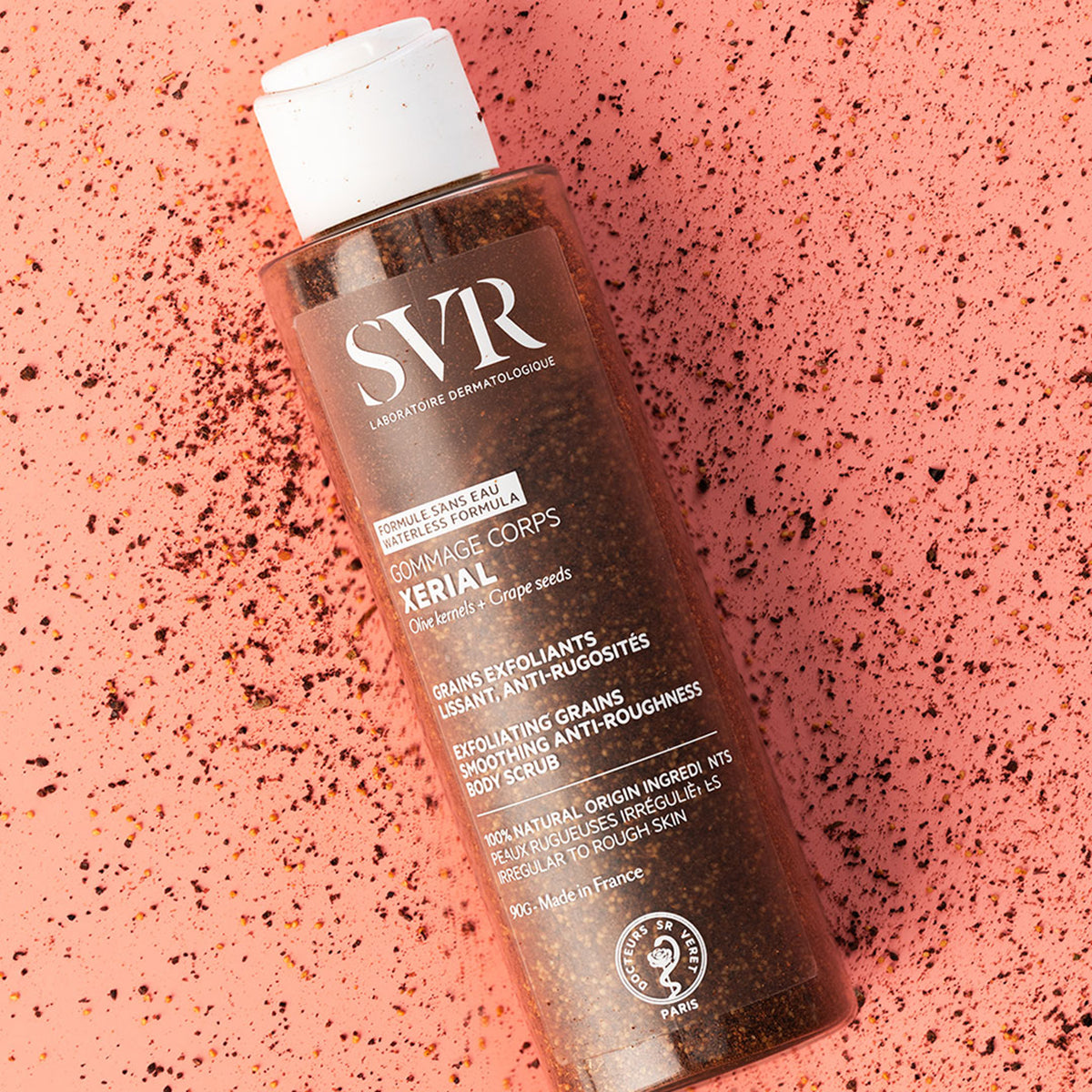 SVR XERIAL Gommage body scrub bottle with exfoliating grains on a textured pink background.