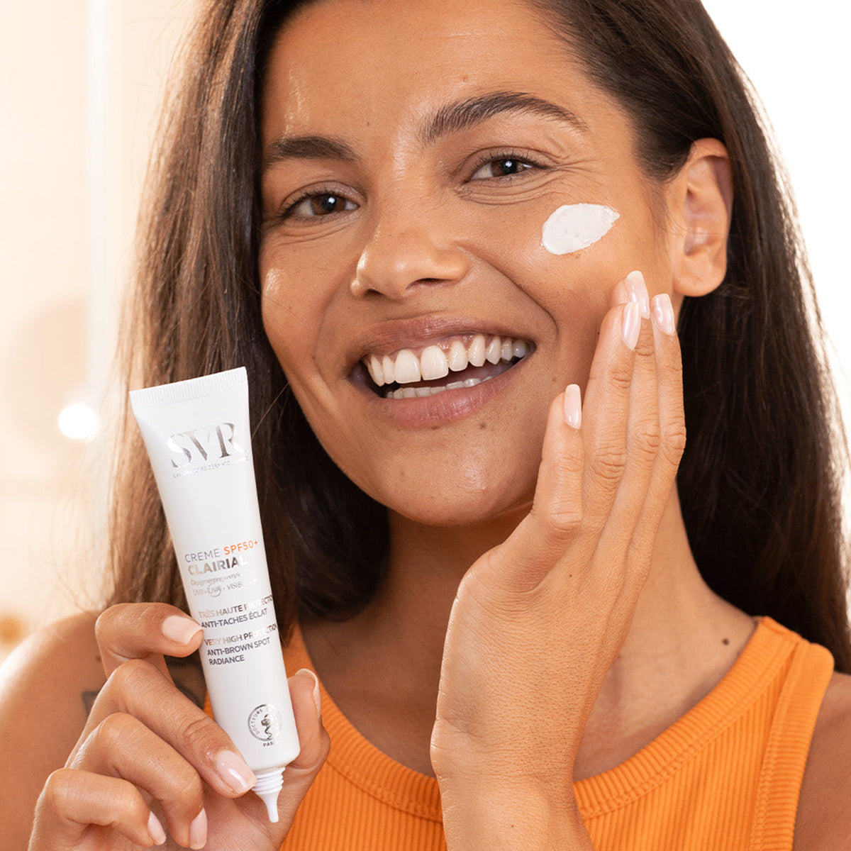 Smiling woman applying white SVR CLAIRIAL Crème SPF50+ to her cheek from a white squeeze tube.