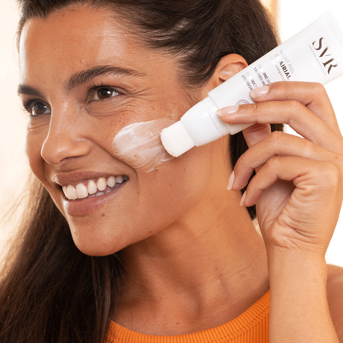 Woman applying SVR Clairial Night Peel from a white squeeze tube with a built-in brush applicator onto her cheek.