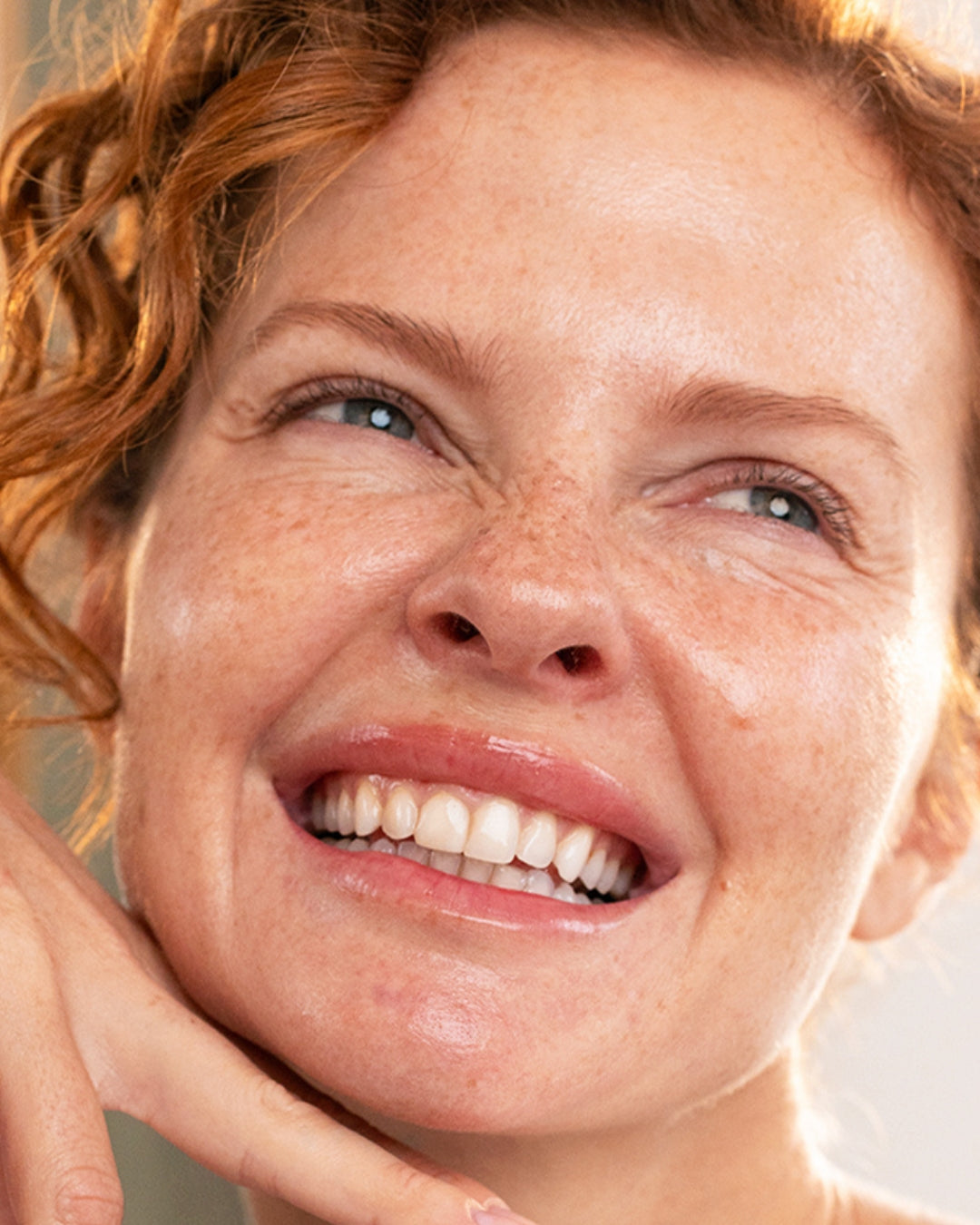 Close-up of a smiling woman with red curly hair and freckled skin, showcasing a natural, radiant complexion.
