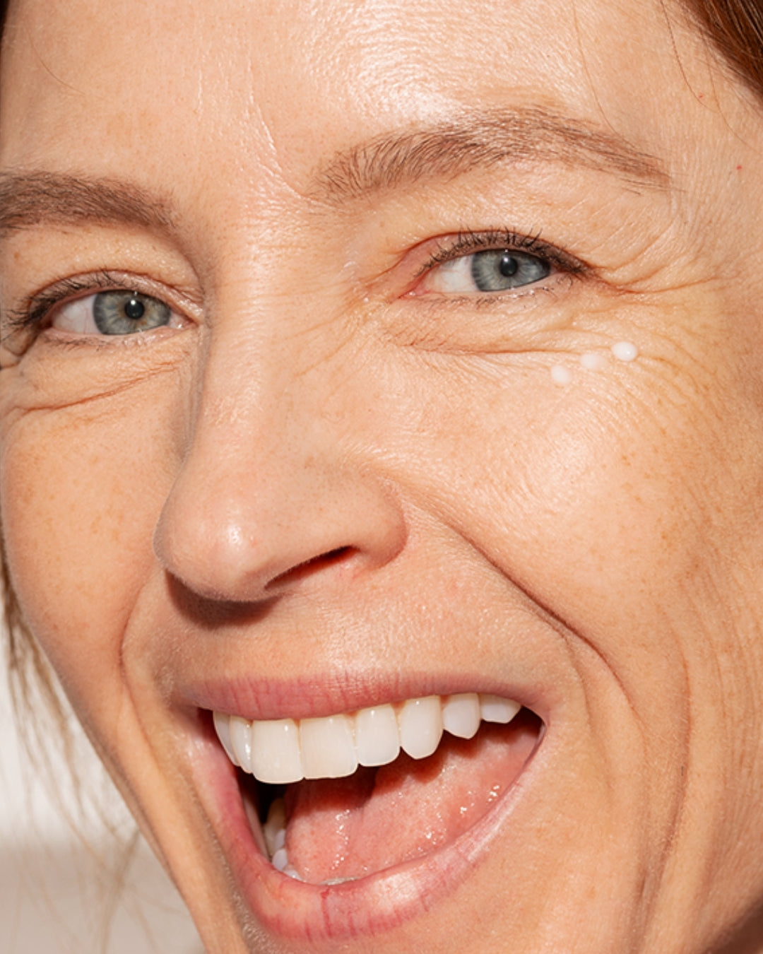 Close-up of a smiling woman with white DENSITIUM eye cream dots applied to the skin under her eye.