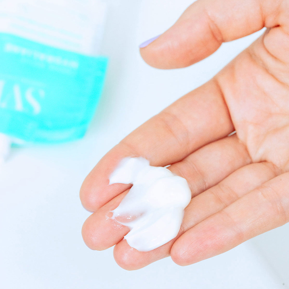 Rich white Hydraliane face cream texture on a palm with a blurred turquoise tube in the background.