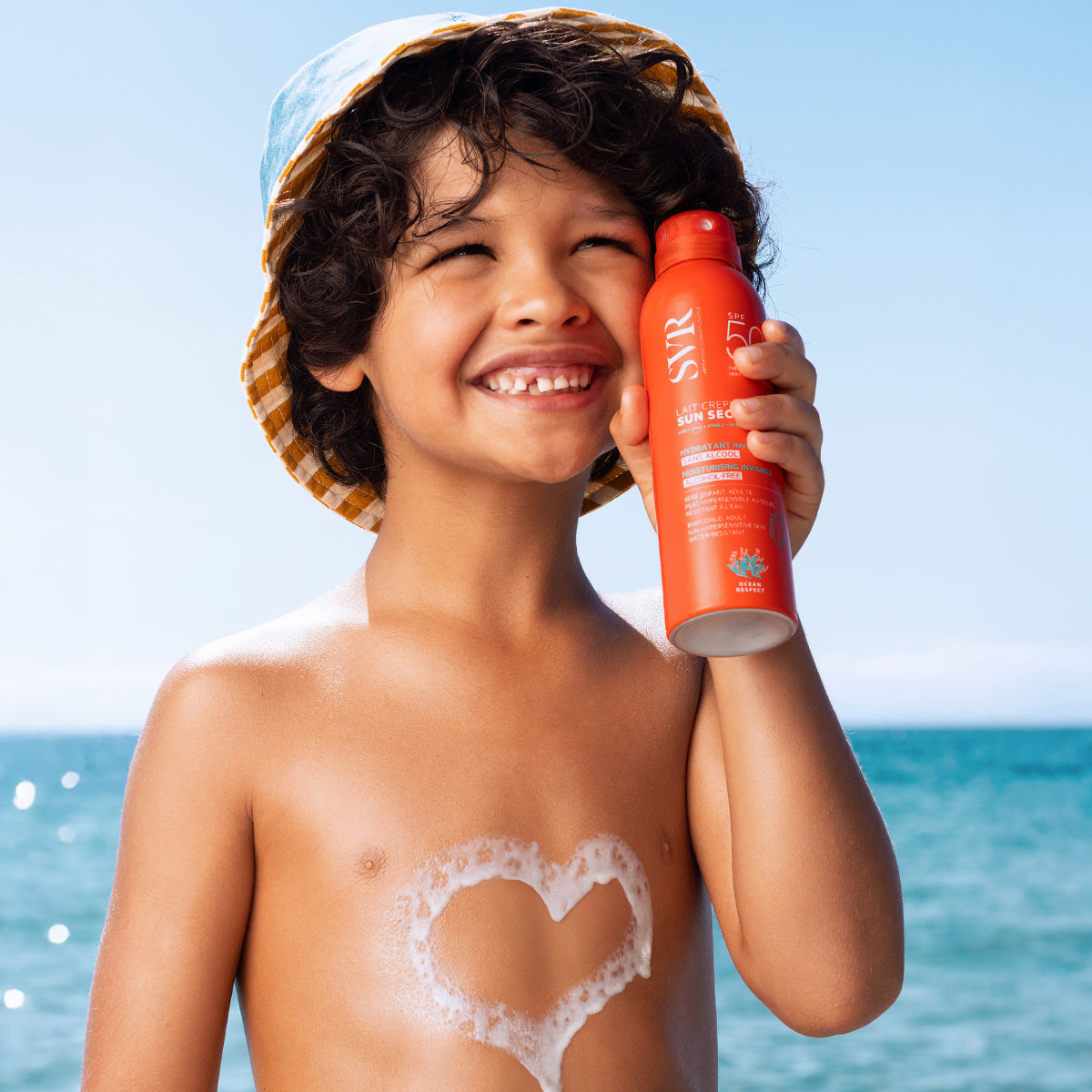 Smiling child in a bucket hat holding an orange SVR SUN SECURE SPF50+ spray bottle with a heart-shaped sunscreen foam on chest.
