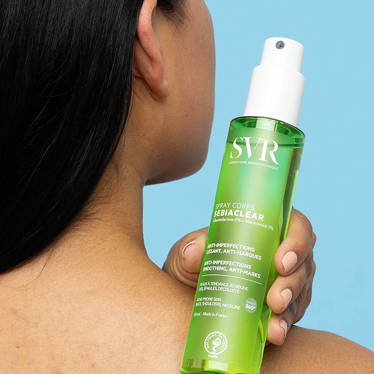 Person holding a green SVR Sebiaclear body spray bottle against their shoulder on a light blue background.