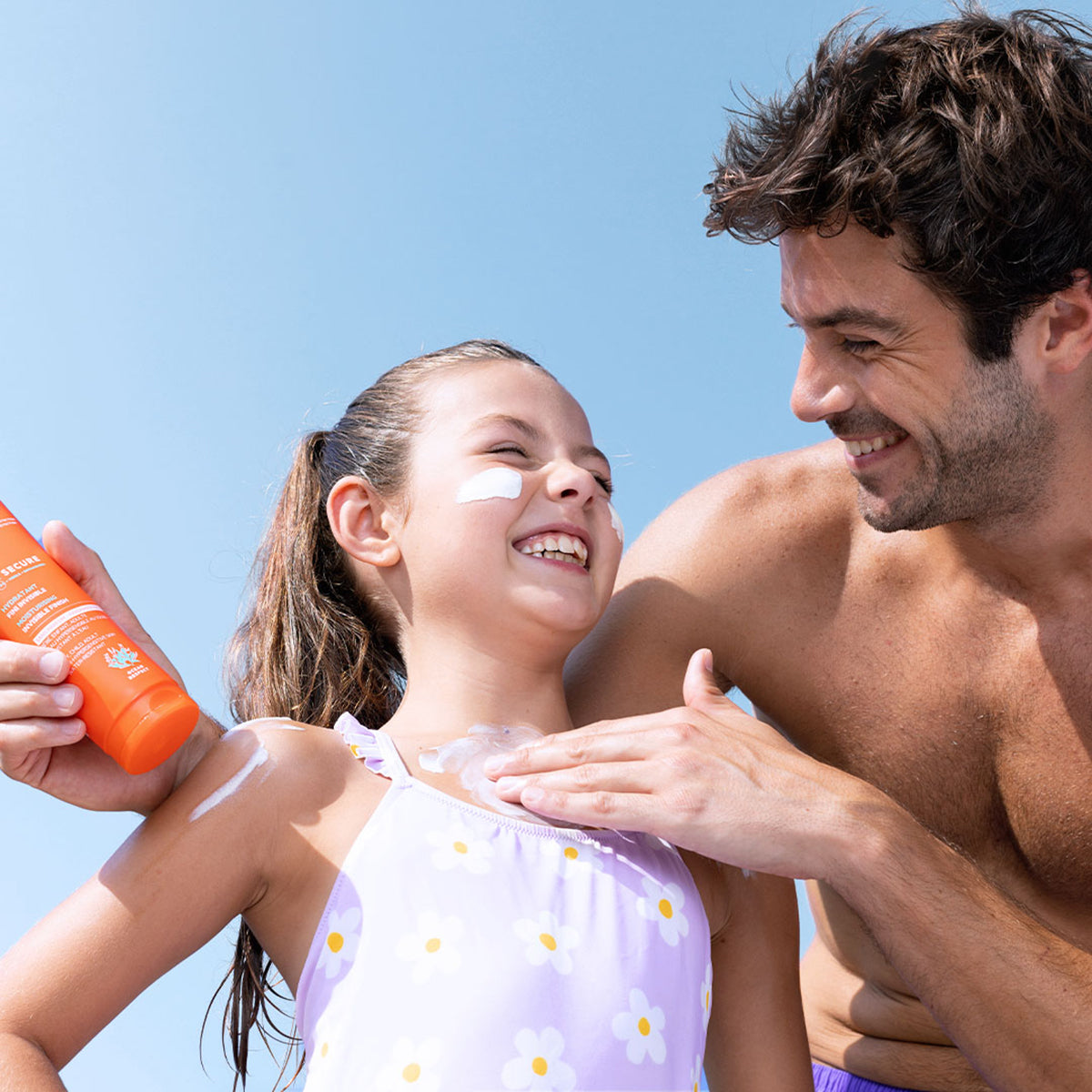 Father applying orange SUN SECURE SPF50+ sunscreen lotion onto a smiling girl wearing a floral swimsuit.