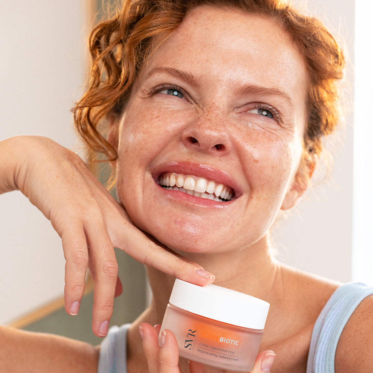Smiling woman holding a 50ml jar of SVR C20 Biotic regenerating radiance cream with a frosted glass finish.