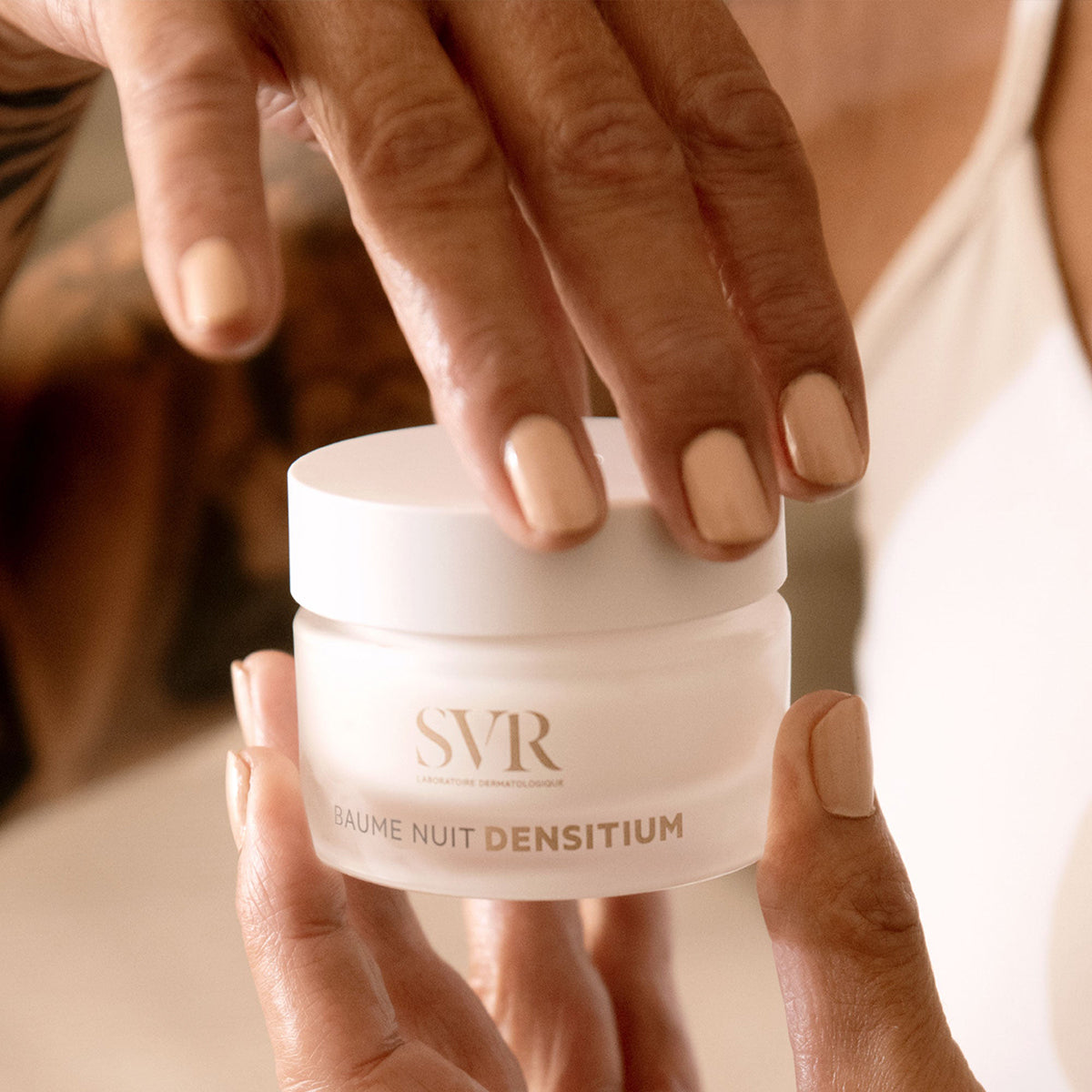 Hands holding a frosted glass jar of SVR Densitium Baume Nuit with a white lid and gold lettering.