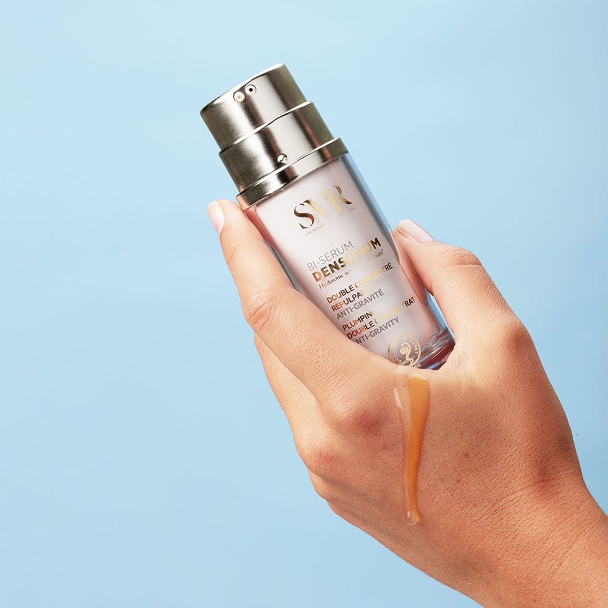 Hand holding SVR Densitium Bi-Serum bottle with a metallic pump, dispensing golden anti-gravity serum onto skin.