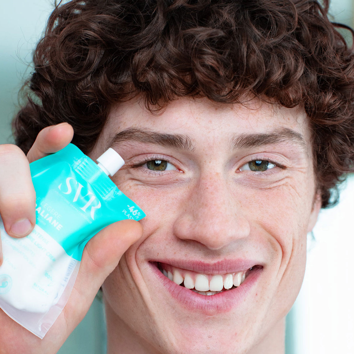 Smiling young man holding a turquoise SVR Hydraliane Crème Légère refill pouch near his face.
