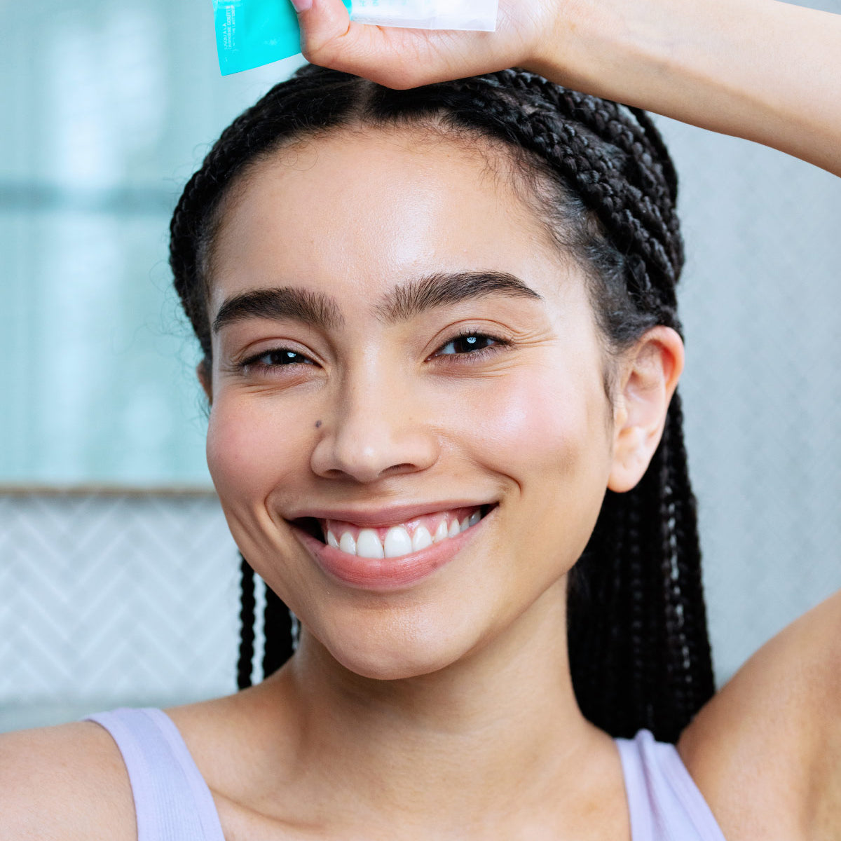 Smiling woman with braided hair holding a teal HYDRALIANE Creme Riche tube against a light bathroom background.
