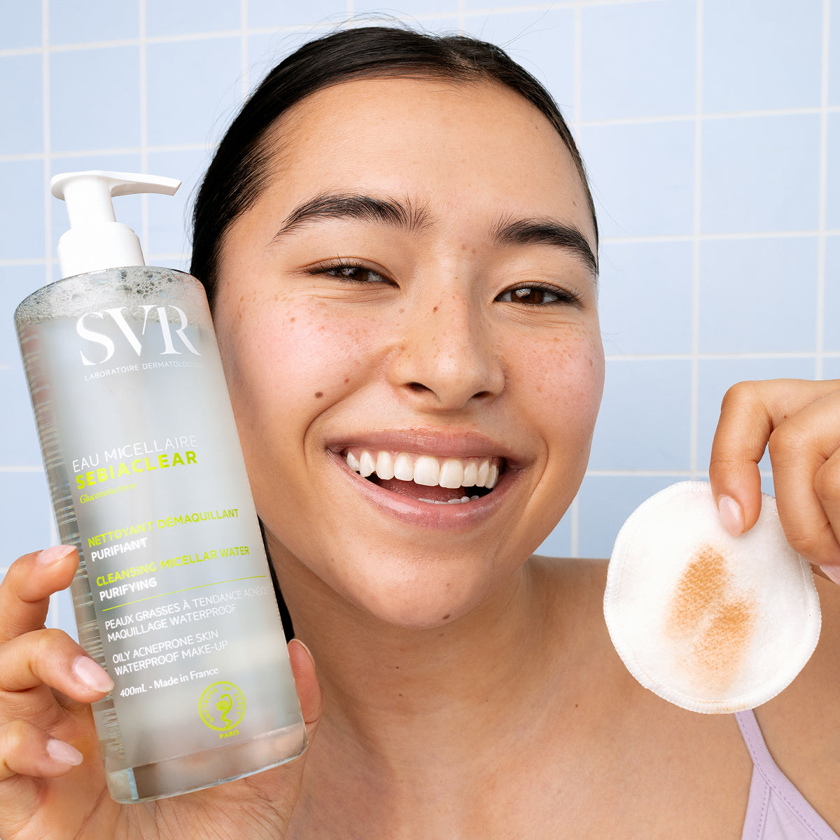 Smiling woman holding a 400ml bottle of SVR Sebiaclear Micellar Water and a used cotton pad against a blue tiled wall.