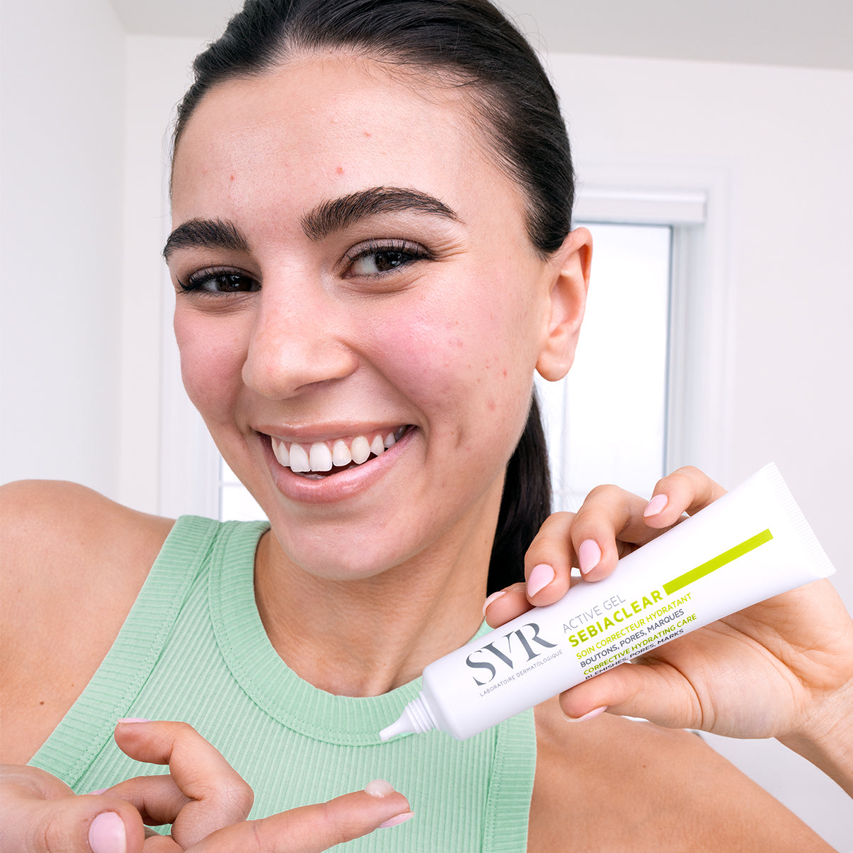 Smiling woman holding a white SVR Sebiaclear Active Gel tube and applying a drop of the clear gel to her fingertip.