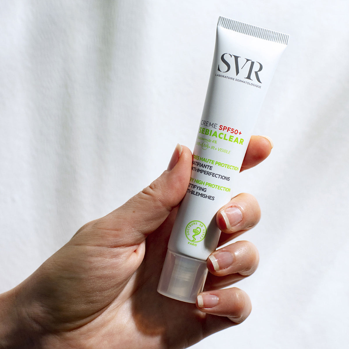 Hand holding a white SVR Sebiaclear SPF50+ cream tube with green and black text against a white background.