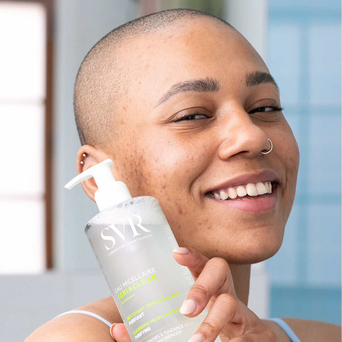 Smiling person holding a clear SVR SEBIACLEAR micellar water bottle with a white pump dispenser.